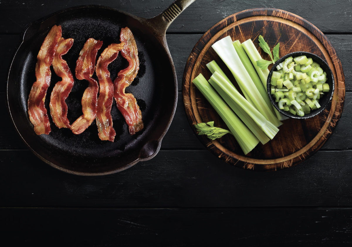 Bacon in skillet and celery stalks next to celery cut up in a black bowl on a wood serving plate.